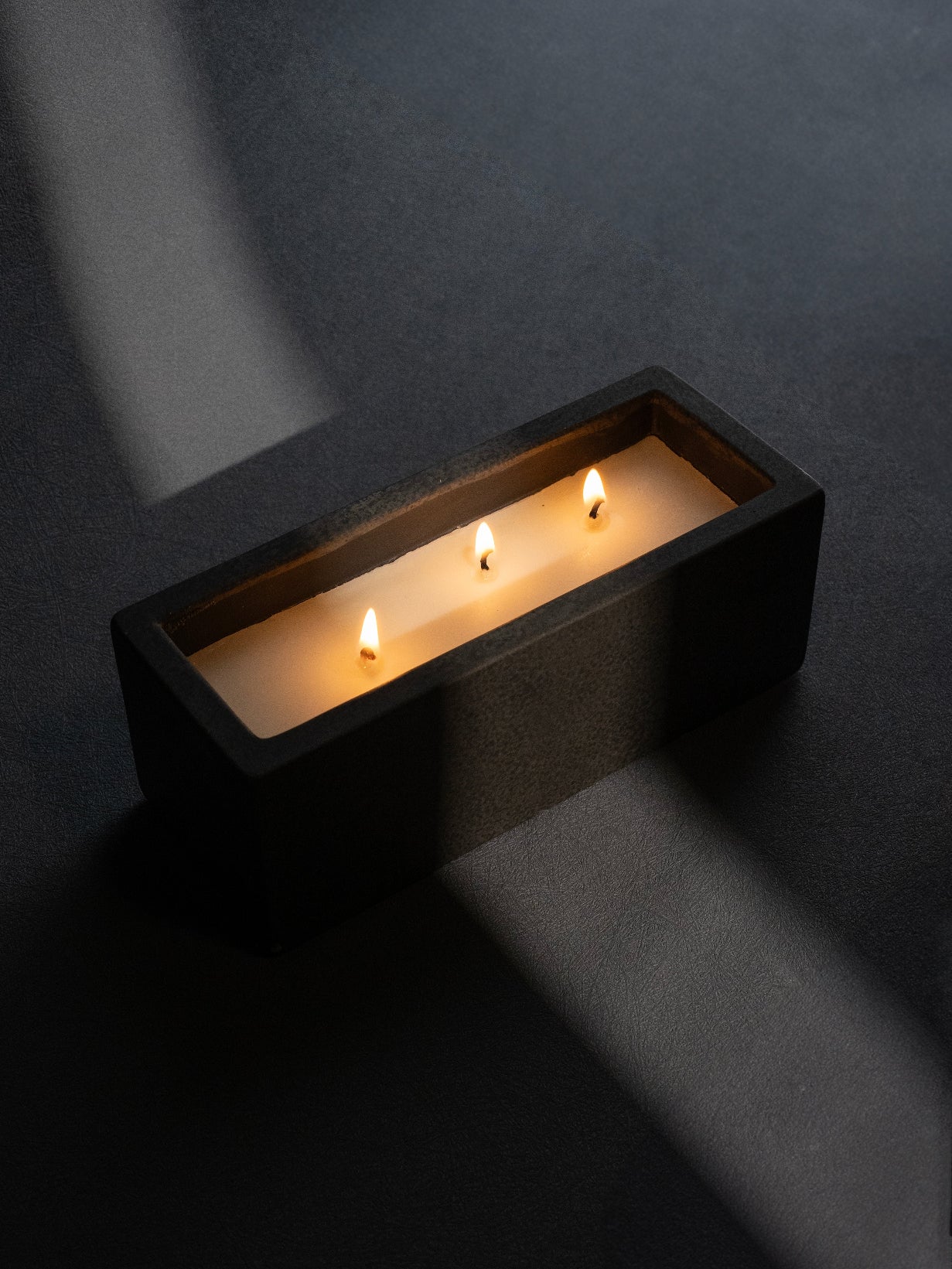A sleek, modern pot with a candle inside, illuminated by its own light, placed on a dark surface with a shadow falling across the scene.